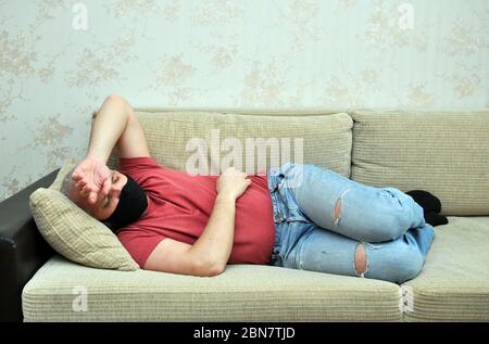 A young man in a black medical mask lies on the sofa with his hand on his forehead. Stock Photo