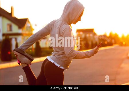 Young female runner, athlete stretching, jogging in the city street in spring sunshine. Beautiful caucasian woman training, listening to music. Concept of sport, healthy lifestyle, movement, activity. Stock Photo