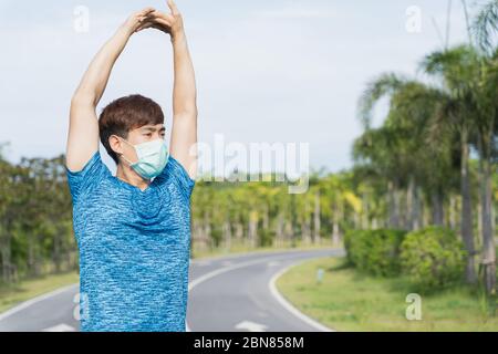 Young male wearing medical mask and stretching his arms before workout training session at the park. Stay in fit during quarantine in the days of the Stock Photo