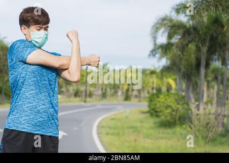 Young male wearing medical mask and stretching his arms before workout training session at the park. Stay in fit during quarantine in the days of the Stock Photo