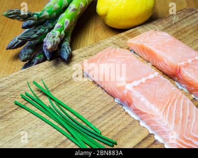 Two raw salmon fillets on a wooden chopping board with fresh chives asparagus and lemon Stock Photo