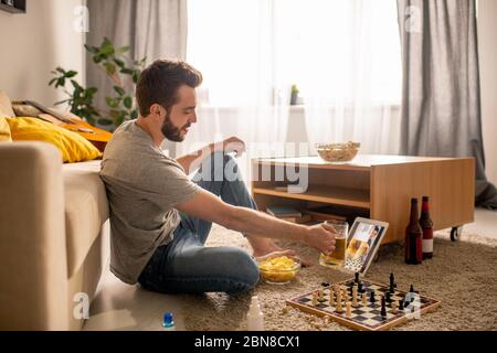 Positive bearded man sitting on floor and drinking beer with friend while communicating online in home isolation Stock Photo