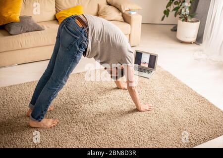 Inflexible young man in jeans practicing downward facing dog pose with trainer online during home isolation Stock Photo