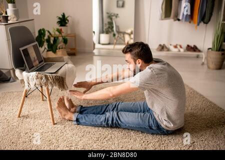 Young bearded man sitting on floor in living room and bending to feet while listening to yoga teacher online Stock Photo