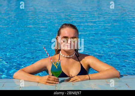 Young european woman holds cocktail drink in blue swimming pool Stock Photo