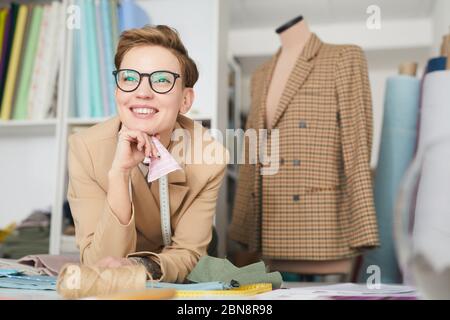 Happy young tailor in eyeglasses working at the table with fabric in the workshop Stock Photo