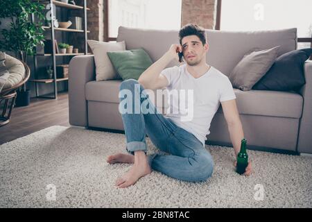 Portrait of his he nice attractive sad depressed guy sitting on carpet talking on phone with friend drinking beer at modern industrial loft brick Stock Photo