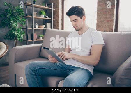 Portrait of his he nice attractive focused brunette guy sitting on divan reading ebook using app web service at modern industrial loft brick interior Stock Photo