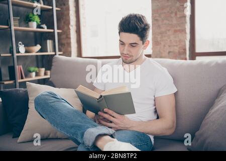 Portrait of his he nice attractive focused peaceful smart clever brunette guy sitting on divan reading paper book at modern industrial loft brick Stock Photo