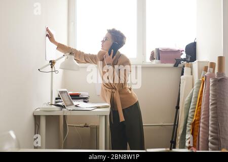 Young designer talking on mobile phone and discussing working moments while working at her office Stock Photo