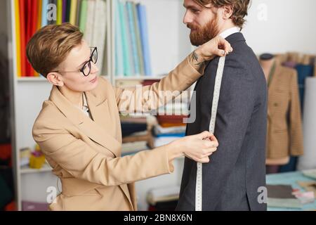Young fashion designer measuring the sleeve of jacket with tape measure while working with her client Stock Photo