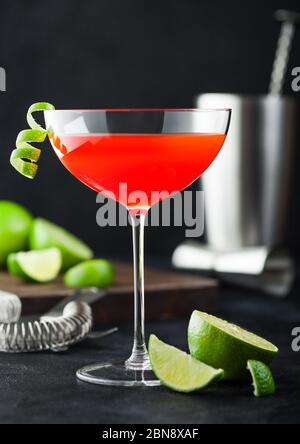 Cosmopolitan cocktail in luxury crystal glass with lime peel and fresh limes with strainer, jigger and shaker on black table background. Stock Photo