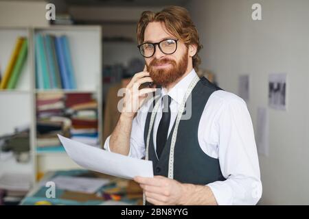 Portrait of young bearded designer in eyeglasses looking at camera while talking on mobile phone with his client Stock Photo