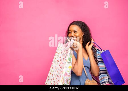 beautiful african lady carrying shopping bags, smiling, and feeling excited on pink background, looking and pointing to the side Stock Photo
