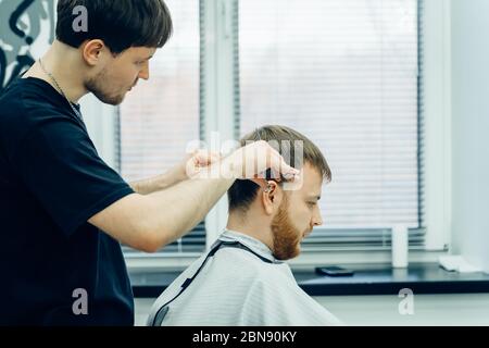 Tattooed Barber cuts the hair of the client with scissors. Close up. Attractive male is getting a modern haircut in barber shop. Stock Photo