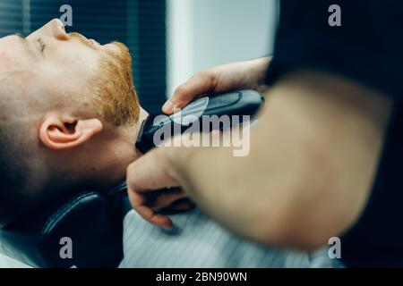 Barber trimming bearded man with shaving machine in barbershop. Hairstyling process. Close-up of a Hairstylist cutting the beard of a bearded male. Stock Photo