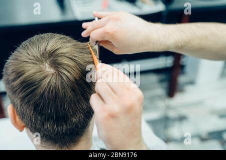 Tattooed Barber cuts the hair of the client with scissors. Close up. Attractive male is getting a modern haircut in barber shop. Close up view. Stock Photo