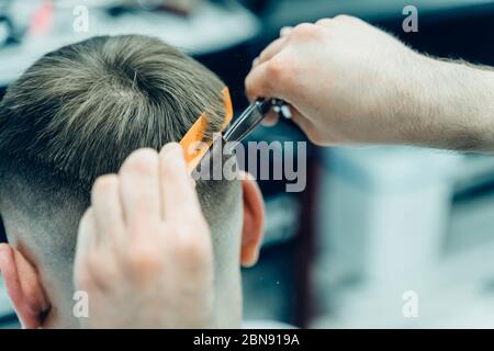 Tattooed Barber cuts the hair of the client with scissors. Close up. Attractive male is getting a modern haircut in barber shop. Close up view. Stock Photo
