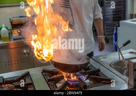 Chef Cooking With Fire In Frying Pan. Professional chef in a commercial kitchen cooking flambe style. Chef frying food in flaming pan on gas hob in co Stock Photo