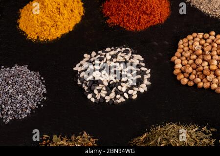 Different piles of spices isolated on black stone plate plaque. Place for text copy space. Various types of condiments directly on a dark stone backgr Stock Photo