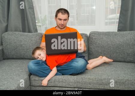 Man in orange shirt tries to work on laptop during his son disturb him. Home office work and stay home concept Stock Photo