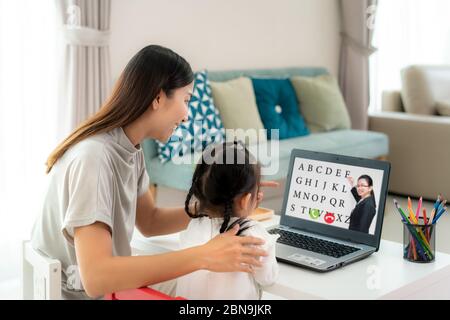 Asian kindergarten school girl with mother video conference e-learning with teacher on laptop in living room at home. Homeschooling and distance learn Stock Photo