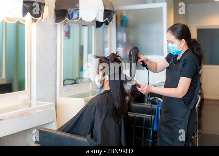 Asian young woman wearing face masks to protect themselves from Covid-19 during hairdresser using Hair dryer and comb to black hair in beauty salon. Stock Photo
