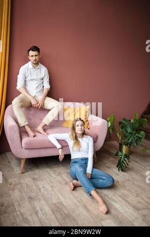 Man sits on pink sofa's back against wall, woman sits on floor near. Stock Photo