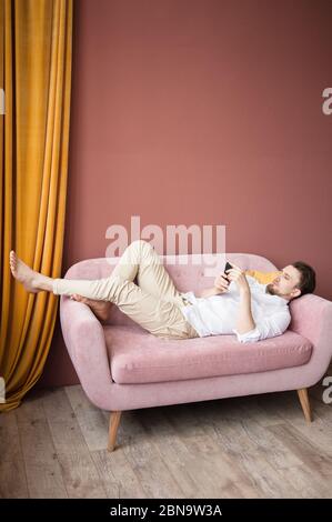 Young man in a white shirt works on his phone while lying on pink sofa Stock Photo