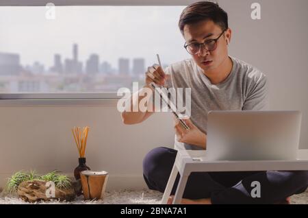 Asian man in casual wear sitting and working on laptop in room that have candle, aroma diffuser and tillandsia airplant on the floor. Concentrated at Stock Photo