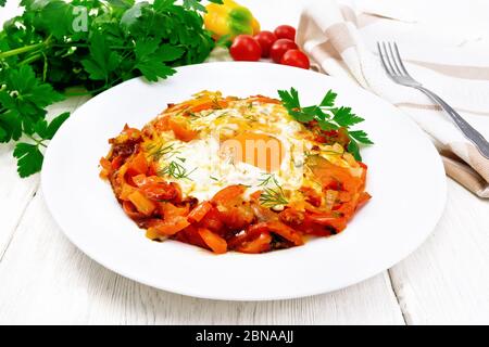 Fried eggs with tomatoes, sweet pepper, onions and herbs in a plate, napkin, parsley and fork on white wooden board background Stock Photo