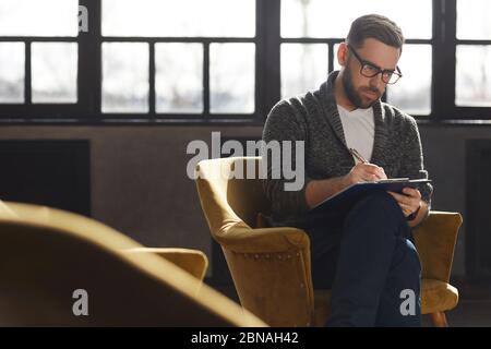 Serious bearded man in eyeglasses sitting on chair and making notes in document at office Stock Photo