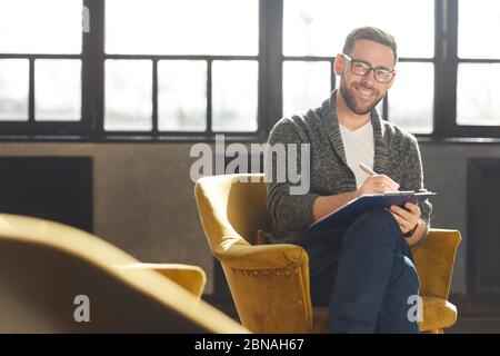 Portrait of bearded young businessman in eyeglasses sitting on chair and smiling at camera while making notes in document Stock Photo