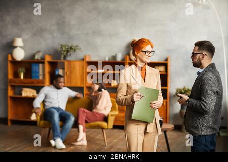 Business colleagues standing and discussing working moments with other people sitting on sofa in the background at office Stock Photo