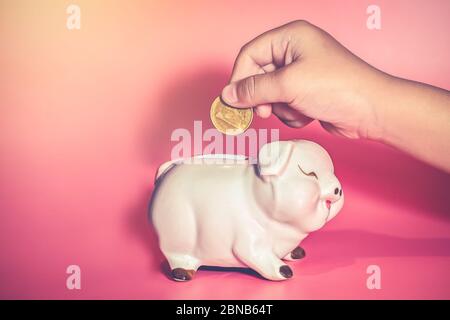 Child's Hand Drop A Coin In Piggy Bank for saving with pile of coins on table at home,Save money for future learning. Stock Photo