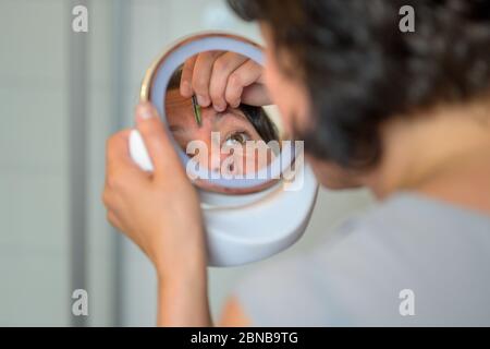 Woman plucking her eyebrows using a small handheld portable mirror in an over the shoulder view with focus to her reflection Stock Photo