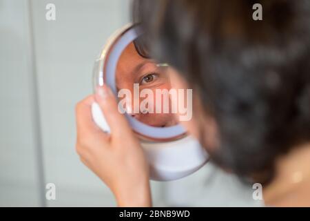 Woman plucking her eyebrows using a small handheld portable mirror in an over the shoulder view with focus to her reflection Stock Photo