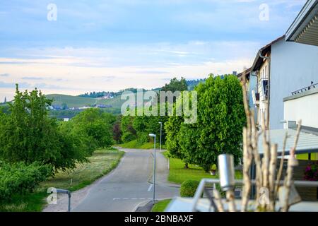 Quiet tree lined rural road in a spring landscape curving past houses on the right towards distant hills Stock Photo