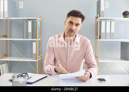 A businessman in a shirt with a document in his hand looks at the camera tells the client in the office. A man in a business office with a contract on Stock Photo