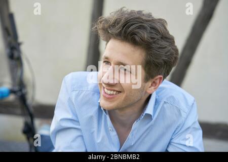 Portrait of a young handsome man with brown curls and a three-day beard, sitting there in a blue stylish shirt, smiling with white teeth Stock Photo