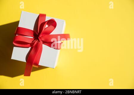 white gift box withred ribbon and bow on yellow background. Copy space.Fashion wrapped square gift or presents box, top view. Flat lay composition for Stock Photo