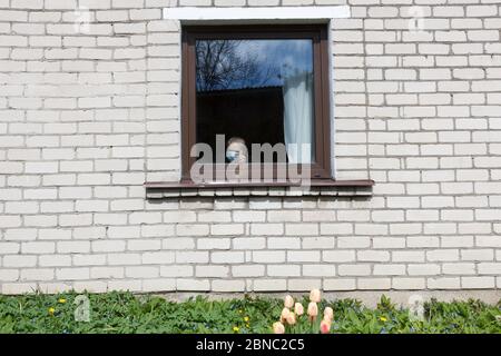 An old grandmother in a protective mask looks out the window at self-isolation. Elderly woman at quarantine at home. Stock Photo