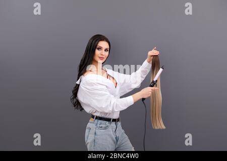 Fashion portrait of female hair artist or stylist holding sections of hair for extension with curling iron on grey background Stock Photo