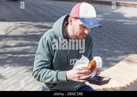 A young man in a red blue and white baseball cap and a khaki sweatshirt with a hood holds in his hands a large juicy burger sitting on the street. A q Stock Photo
