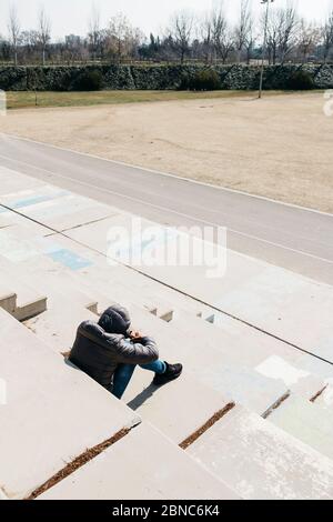 a young caucasian man, seen from behind, wearing jeans and a gray hooded jacket, curled up sitting on an outdoor concrete stairway Stock Photo