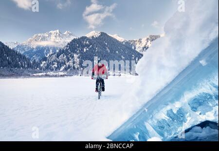 Man in red jacket is riding his bicycle at frozen lake with blue ice at foreground at snowy mountains at background Stock Photo