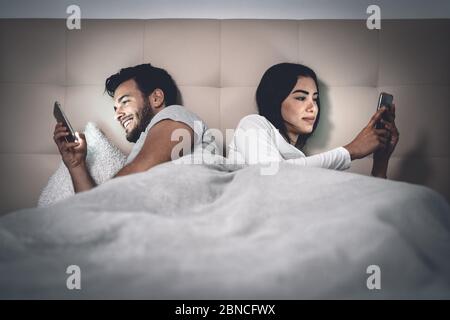 Young couple lying in bed using mobile phone while ignoring each other - Man and woman addicted to smartphone social networks technology