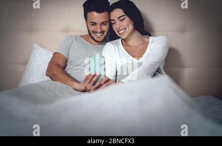 Young couple using mobile phone in bed - Man and woman watching smartphone and having fun with social networks technology apps Stock Photo