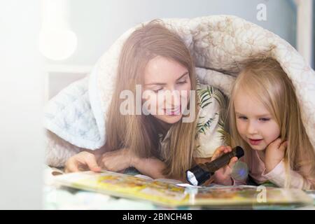 Closeup portrait of young mother reading a book using flashlight with her happy 3 years old daughter Stock Photo