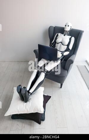Woman robot working with laptop while sitting in cozy armchair putting her legs on pillow Stock Photo
